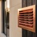 Close-up of a patinated copper dryer vent termination with a louvered grille on a modern limestone and charred cedar exterior, side-lit at golden hour, with shallow depth of field and blurred window mullions and landscaping in the background; slight angled view shows a hint of rigid metal ducting inside.