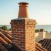 Low-angle photo of a historic red-clay brick chimney with a terracotta pot on a Mediterranean Revival clay-tile roof in Tampa Bay at golden hour, with palm fronds, stucco, and a glimpse of the bay softly blurred; subtle Spanish Colonial and Victorian rooflines in the distance.